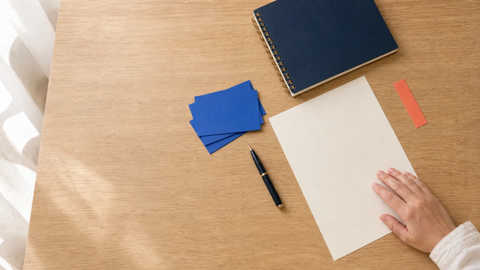 Blank study paper, blue cards, navy notebook, and a resting hand on a warm desk